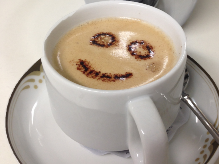 Tasse mit Kaffee, in dem ein lächelndes Gesicht aus Schaum gemalt ist.
