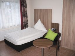 Ein einfaches Zimmer mit einem Einzelbett, Tisch und Stuhl. Fenster mit Vorhängen.