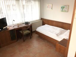 Helles Zimmer mit Einzelbett, Schreibtisch, Stuhl und Fernseher.